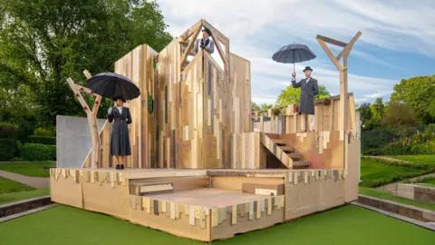 Colin J Smith A theatre set in a wooden raised platform design with three people standing on it. They are wearing dark jackets and hats and two are carrying black umbrellas.