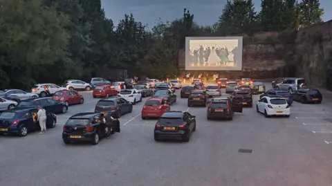 Karen Hall Cars are parked facing a large outdoor cinema screen in the evening. 