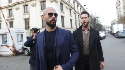 EPA The Tate brothers appearing at court. Andrew is in the front with a bald head and sunglasses, and his brother Tristan is standing behind him