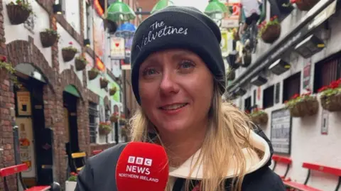 Leanne is smiling into the camera. She is dressed in a white hoody, navy coat and black beanie hat while holding a red microphone. 