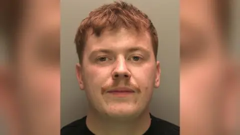 A police custody image of Brandon Barker. He has short, cropped red hair, freckled skin and a small moustache. He is staring into the camera and is wearing a black t-shirt.