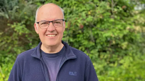 Jeremy Norton, wearing T-shirt and casual top, smiling at the camera