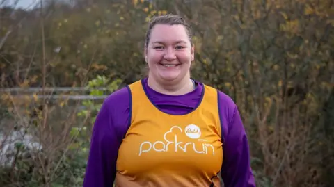 Alannah Mathewson is a woman in her 20s wearing a purple top and an orange vest with the Parkrun logo in white. She is standing in front of some trees with a river behind 