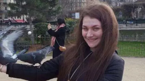 Humberside Police Libby, who has long brown hair and wearing a black coat, with two pigeons on her right arm standing in a gravel and lawned park area