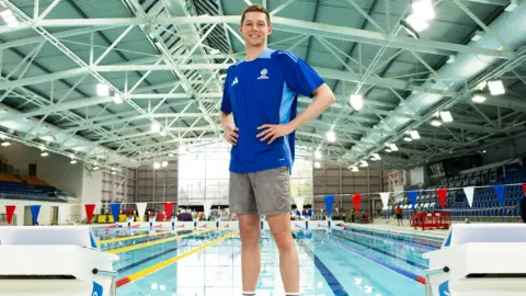 Duncan Scott standing in front of a swimming pool. He is wearing a blue T-short and grey shorts. There is bunting across the pool behind him.