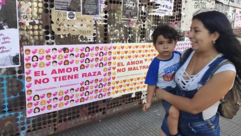 BBC A woman with a child walks past a wall with posters saying "love does not have a race"