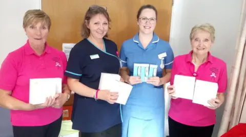 Royal Walsall NHS Healthcare Trust Four women stand in a row smiling as they hold up white boxes. The women on each end wear bright pink t-shirts with a pink breast cancer ribbon printed on the breast. The two in the centre are wearing blue nurse uniforms. they are standing in a hospital ward