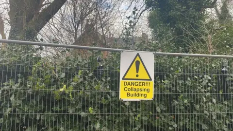 A sign which reads 'Danger!!! Collapsing Building' on the fence surrounding the building.