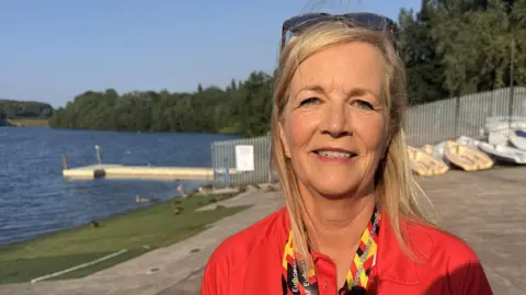 Kathy Yates, who is wearing a red T-shirt and has two medals around her neck, is smiling at the camera in front of the reservoir. 