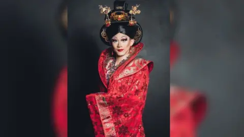 Zee A person in a bright red rob with a black wig and elaborate makeup