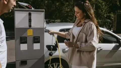 Getty Images Smiling mother holding electric plug standing by father at gas station