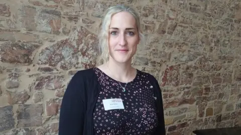 Anna Tarrant A photo of professor Anna Tarrant stood in front of an old-fashioned looking stone wall. She has light blonde hair and is wearing a black floral top with a black cardigan. She has a silver necklace on and a white name sticker on her top that we can't make out the wording on.