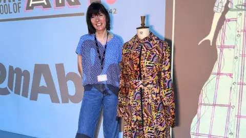 BBC/ Emily Johnson A woman stands beside a dress form displaying a brightly coloured patterned dress with a collar and belt, set against a projected backdrop showing text and a fashion sketch. The woman wears a blue patterned shirt, jeans and a lanyard.