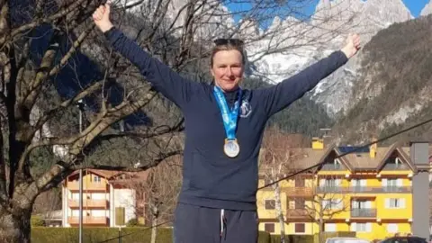 Helen Hislop is smiling, with her arms raised in the air, as she stands on a podium while wearing her gold medal. 