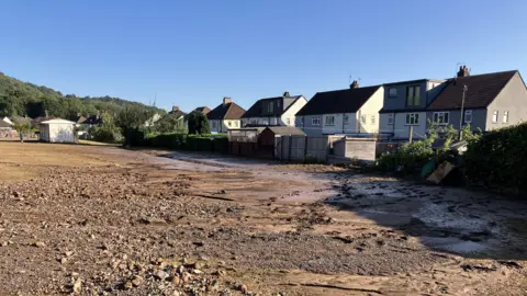 A football pitch which is covered in wet mud and stone sits with houses on Heol Berry backing on to it.
