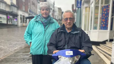 Julia Gregory / BBC A man, sat on a blue mobility scooter, and a woman stood to his left. They are looking at the camera. They are in a rainy street with cobbled stones and shops behind them.
