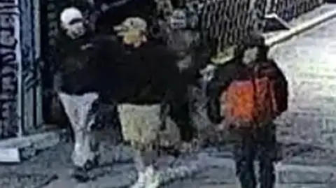A grainy CCTV screenshot that shows four men walking over a bridge. It is hard to make out any details but one wears a black and red jacket with the hood up