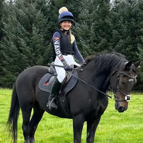 Katie Nairn A young girl sitting in a saddle on a dark-coloured horse. She is wearing a riding helmet and smiling at the camera. 