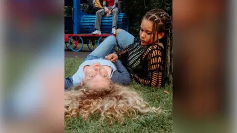 Rooscreen Amelia Clarkson with long curly blond hair wearing a blue top and jeans and lying on the grass of a playground. Ebony Aboagye has locs and is wearing a dark-coloured top. She is lying on the grass beside Amelia. A pair of legs in jeans sitting on a toy train is visible in the background