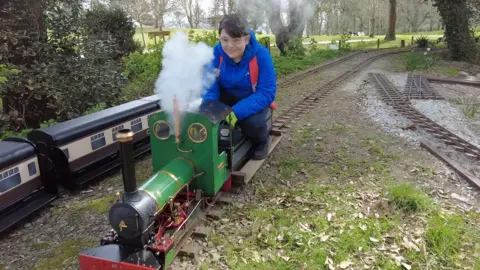 Harley, wearing a blue jumper, rides a green model steam engine. Steam is puffing out of the top of the funnel.