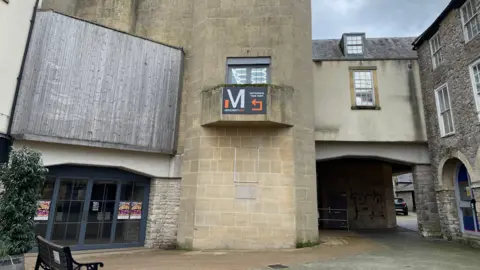 BBC The picture shows the outside of the Amulet Theatre in Shepton Mallet. There are stone walls and wooden panels.  There is a bench to the left of the picture,