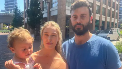 Baby Sienna, Sarah and Jon stand outdoors facing the camera in bright sunlight outside modern brick buildings and parked vehicles.