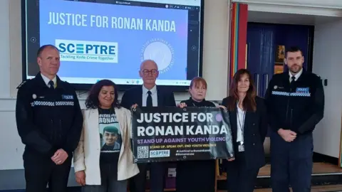 A group of people standing infront of a class of students holding a sign about knife crime and justice for Ronan kanda