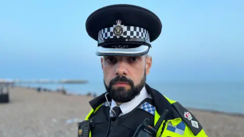 Charlie Rose / BBC A police officer wearing a hi-vis jacket and formal peaked police hat. He is stood on a stony beach with the sea in the background.