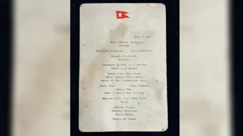 Henry Aldridge & Son Ltd First-Class menu from the Titanic. It has water stains on it and a red emblem of a flag at the top.