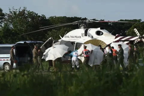 Lillian Suwanrumpha / AFP Police and military personnel use umbrellas to cover around a stretcher near a helicopter and an ambulance at a military airport in Chiang Rai