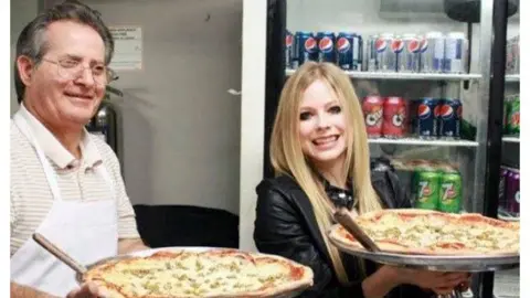 Avril Lavigne / Facebook Lavigne serves a slice of her signature pizza with restaurant owner Bill Kosmopoulos