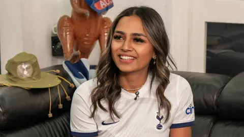 @switchedonsef Davina, a South Asian woman smiling looking to the right of the camera. She is wearing a white Tottenham Hotspur jersey. Behind her is a green hat and inflatable brown animals.