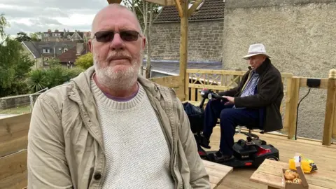 Mike Somerton sitting in the rebuilt shed, with a man on a mobility scooter behind him