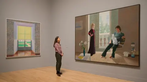 AFP A woman stands between two David Hockney paintings - Contrejour in the French Style 1974, left, and Mr and Mrs Clark and Percy. The first painting depicts a window, surrounded by floral wallpaper, giving a glimpse of pathways and manicured grass from beneath a yellow window blind. The second depicts Ossie Clark, sitting facing the viewer with a white cat on his lap, and his wife Celia Birtwell, standing with her hand on her hip.