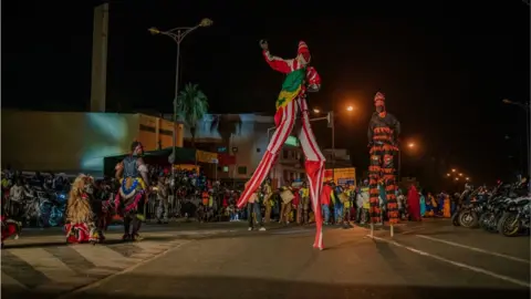 AFP Two stilt walkers wearing colourful clothing with a crowd behind them