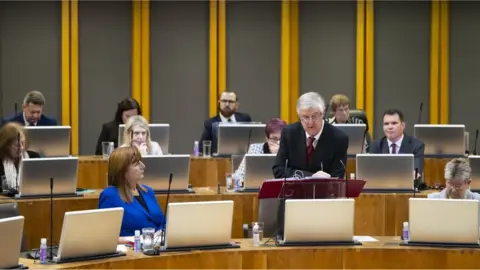 Senedd Cymru View of a group of Labour Senedd members, including Mark Drakeford, who is stood up making a speech.