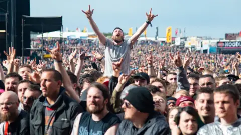 Getty Images Festival goers cheering in the crowd during day 2 of Download Festival 2019. A white man with a beard sits above the crowd on another spectator's shoulders, his hands stretched up making the sign of horns. The hand gesture, associated with rock music, is repeated by other members of the crowd. The audience is outside on a sunny day and in the background flags and food outlets can be seen.