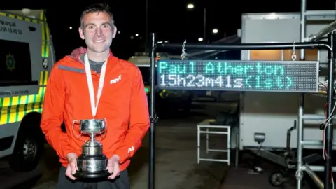 Manx Radio Paul Atherton holding the trophy with his finishing time on a board behind him