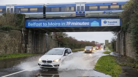 PA Media A car moves through a deep water on a road near Moira in County Down as a train crosses a bridge overhead
