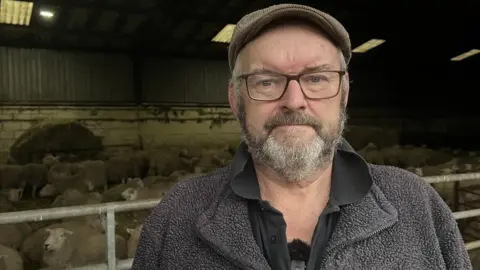 BBC Dave Williams on a farmyard with sheep behind him in a barn