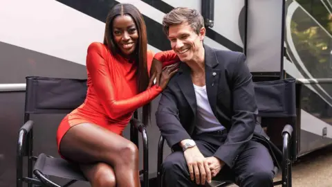 Kieron McCarron / ITV AJ Odudu, wearing a red dress, and Big Brother co-host Will Best, wearing a black suit, sit in black director-style chairs, smiling