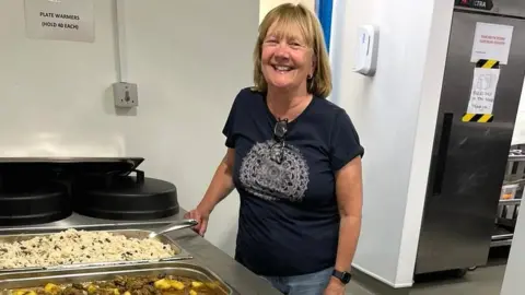 Julian Trust Jane standing by trays of hot food in a kitchen. She is wearing a blue T-shirt and jeans and smiling