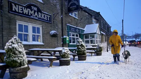 PA Media/Owen Humphreys A woman in a yellow coat walks a dog along a snow covered path next to a pub