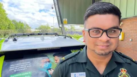 male paramedic in front of a rapid response vehicle which has an anti-violence campaign picture of him in the window