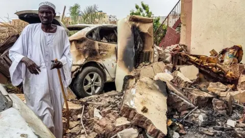 Getty Images A man inspects damage as he walks through the rubble by a destroyed car outside a house that was hit by an artillery shell in the Azhari district in the south of Khartoum on June 6, 2023