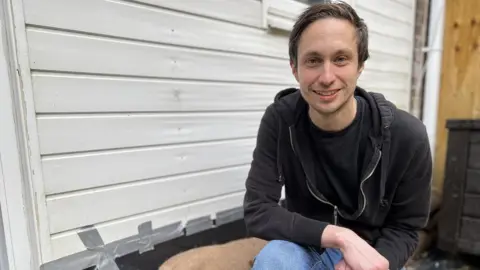 BBC Paul Stoner crouches in front of his garage door in front of his newly delivered sandbags