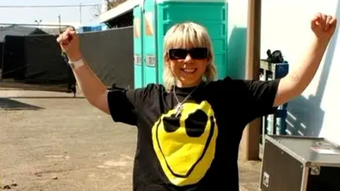 Meg Meg is in front of some portable toilets in a backstage area of a festival on a bright sunny day. She's wearing an oversized black t-shirt with a yellow smiley face on it. She's got her arms in the air and she's smiling. Her blonde fringe touches the top of her large black sunglasses