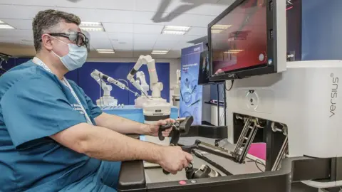 MIKE DEAN/EYE IMAGERY Consultant urologist Mohamed Abdulmajed at the controls of the surgical robot, which is operating in the background behind him