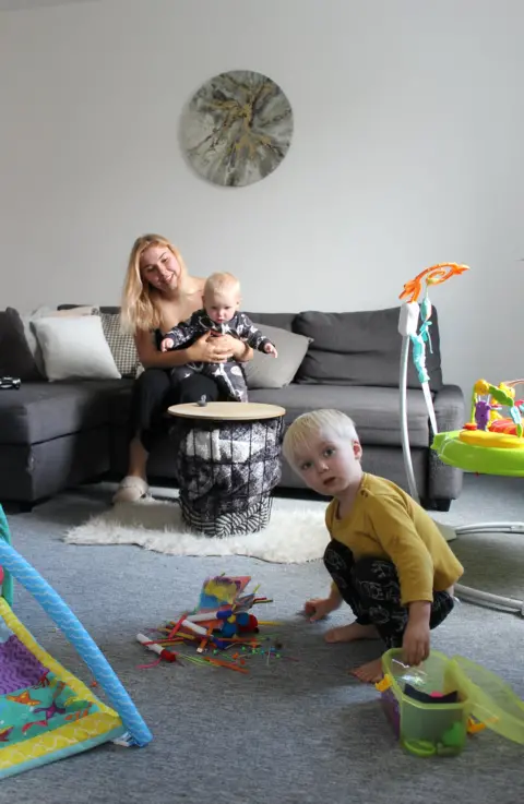 BBC A mother sits on the sofa holding her baby as her toddler plays with his toys on the floor