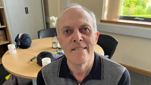 Ollie Conopo/BBC David Holmes - man with short white hair and blue shirt sitting in an office